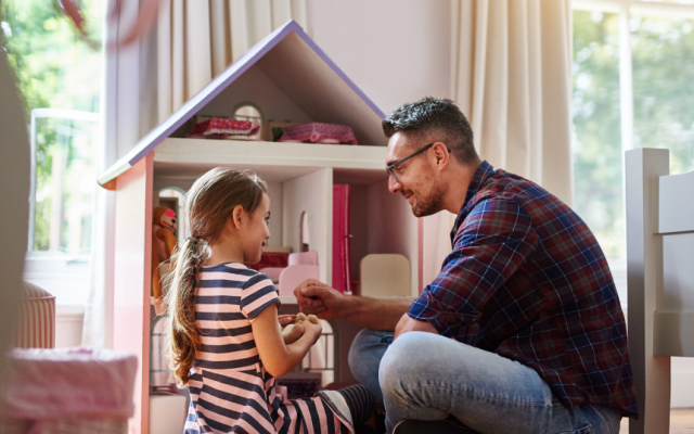 Primary school aged girl and man wearing glasses sit in front of doll house chatting representing fostering family after foster care approval